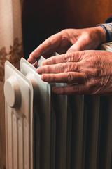 An elderly man warms his hands over an electric heater. In the off-season, central heating is delayed, so people have to buy additional heaters to keep houses warm despite increased electricity bills