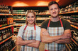 © estradaanton - Professional young man and woman stand in grocery store and pose on camera. She hold hands crossed and smile. Happy positive workers wear apron.