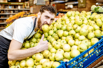 Wall Mural - Funny portrait of a handsome shop worker hugging a pile of apples in the supermarket