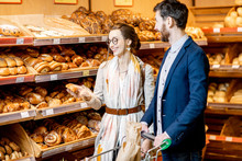 Young And Happy Couple Choosing Fresh Pastries, Standing Together With Shopping Cart In The Bakery Department Of The Supermarket