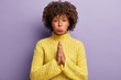 Unhappy woman pleads for forgiveness, keeps palms together, has regretful facial expression, believs God will help you to achieve desirable wish, wears yellow sweater, models over purple wall