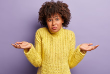 Doubtful Confused Afro American Young Woman Spreads Hands In Both Sides, Makes Decision About Future Plans, Has No Make Up, Curly Hair, Wears Yellow Winter Sweater, Isolated On Purple Background