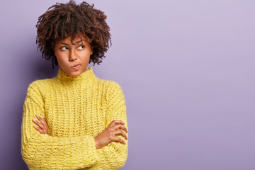 Canvas Print - Hmm let me think. Thoughtful dark skinned woman with curly haircut, purses lips, keeps arms folded, wears yellow jumper, makes decision in mind, stands over purple background, mock up space.
