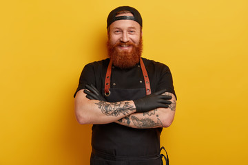 Unshaven smiling red haired man in black uniform, glad to see many visitors in restaurant, keeps tattooed hands crossed, has ginger beard, likes his profession, isolated over yellow background