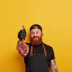 Wall Mural - Attractive unshaven foxy skilled male meat cutter holds fresh meat, wears black cap and apron, waits on customer in retail store, prepares special cuts upon request, stands over yellow background.