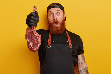 Wall Mural - Surprised cheerful male butcher holds fresh raw piece of meat with bone, has red thick beard, dressed in special working uniform, prepares for making sausages or minced meat. Butchering concept