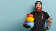 © WHstudio Leushin N - Image of smiling pleased chef holds hand on waist, carries utensils, wears apron and t shirt, being in high spirit for cooking something delicious, stands against blue wall with free space aside