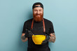 Baking and cooking concept. Happy ginger male chef holds bowl and whipping utensils, whisks eggs in yellow container, wears black uniform, has glad look at camera, isolated on blue background
