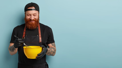 Wall Mural - Satisfied chef mixes ingredients in bowl with whisk, holds kitchenware, prepares delicious dish, wears rubber gloves, black uniform, poses over blue background with empty space for your promotion