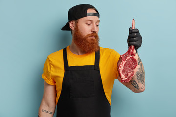 Wall Mural - Photo of serious red haired bearded male chef holds piece of meat, going to prepare pork on grill, shares recipe with colleague, wears uniform and protective rubber gloves for maintaing hygiene