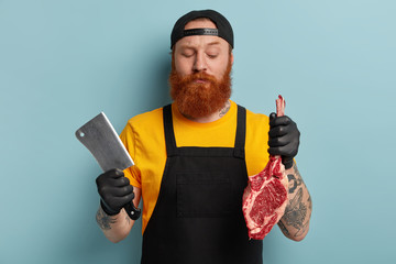 Wall Mural - Waist up shot of serious ginger male butcher seperates meat from bone, holds raw piece of meat, wears rubber protective gloves and apron, transforms beef to supermarket, isolated on blue wall