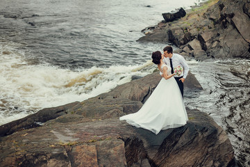 The bride in a luxurious white dress and a beautiful groom hugging against the background of nature. Newlyweds are on a rocky shore, around them the water is boiling