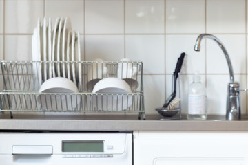 Blur kitchen background with dishwashing machine, water tap, sink, dish rack and plates cups on it, horizontal