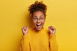 Monochrome shot of overjoyed dark skinned woman with combed Afro hair, smiles broadly, keeps fists clenched, rejoices having triumph, dressed in yellow sweater, round spectacles, celebrates something