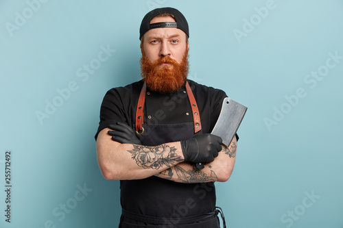 Serious good looking red haired meat cutter holds sharp knife or ceaver in hand, keeps arms folded, wears black special unform, ready for work, poses against blue background. Manufacturing job