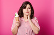 © sementsova321 - Attractive lady looks at camera, holds fresh milk ice cream, keeps hand on cheek, has toothache. Brunette sufers from sensitive tooth disease, eats cold sundae, stands on pink wall background.