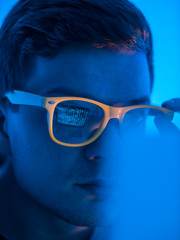 Close-up: A young hacker guy in yellow glasses is working on a cyber-attack computer, while the green symbols of a binary code are reflected in the glasses of his glasses. Programmer surprised