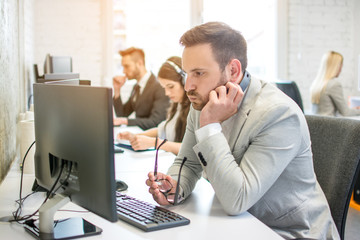 Wall Mural - Confused business man with headset and eyeglasses in hand looking at desktop computer screen while working at his workplace in office