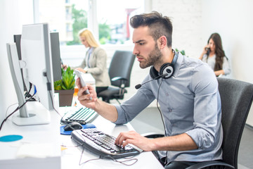 Wall Mural - Side view portrait of handsome young man with headphones around his neck reading something on smart phone while working on computer in office.