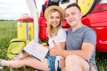 Wall Mural - Young couple traveling in a red car stopped for camping and exploring the route map further way sitting on a green lawn