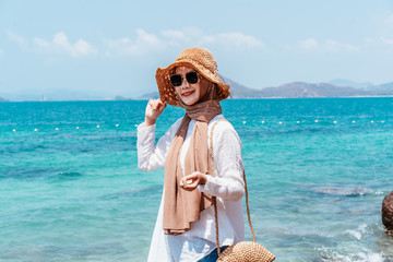 Wall Mural - Young confident muslim woman white dress catching a hat on seashore. Travel Vacation Retirement Lifestyle Concept. asian woman wear sun glass looking on the beach in vacation day. summer time.