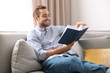 Handsome young man reading book on sofa at home