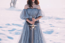 Warlike Girl With Dark Hair In Long Gray Vintage Light Dress, Lady Of Cold And Frost, Bare Open Shoulders And Sharp Silver Sword In Hands, Goddess Of Death With Red Lips, Character For Computer Games