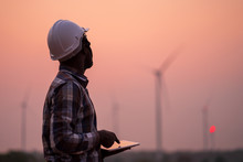 African Engineer Wearing White Hard Hat Standing With Digital Tablet Against Wind Turbine On Sunny Day