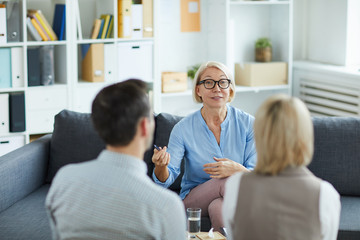 Wall Mural - Mature blonde female psychologist sitting in front of young troubled couple and consulting them in office