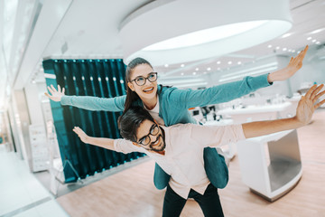 Canvas Print - Cheerful multicultural couple with eyeglasses dressed casual having piggyback at tech store.