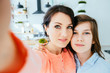 © Iryna - Mother and son at home in kitchen together taking selfie pictures on phone hugging parent to son smiling cheerful looking camera close-up.