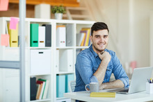 Happy young successful office worker in casualwear sitting by desk in front of camera and networking