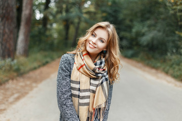 Pretty happy young woman with a stylish hairstyle with a beautiful smile in a spring stylish coat in a fashionable beige vintage scarf walks to the park. Attractive elegant girl relaxes outdoors.