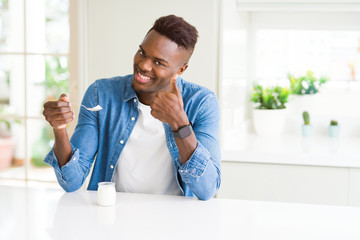 Wall Mural - African american man eating healthy natural yogurt with a spoon happy with big smile doing ok sign, thumb up with fingers, excellent sign