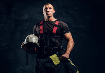 Wall Mural - Portrait of a brutal muscular fireman holding a helmet and jacket standing in the studio against a dark textured wall