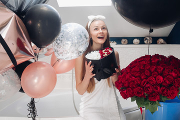 Wall Mural - Young girl laughing and enjoying beautiful presents at home. Happy woman sitting neat big balloons and big bouque of red roses, keeping heart of flowers and posing. Concept of happiness and love.