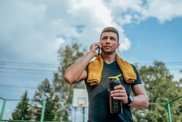 Wall Mural - Athlete man stands summer sports ground city. In his hand bottle of water, resting after exercise. Calls by phone. Trainer guy in watch tracker bracelet after jogging. Workout lifestyle. Free space.