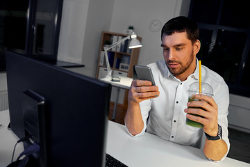 Wall Mural - business, overwork, deadline and people concept - businessman using smartphone and drinking green smoothie at night office