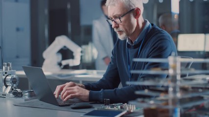 Poster - Professional Electronics Design Engineer Wearing Glasses Works on Laptop Computer in Research Laboratory. In the Background Motherboards, Circuit Board, Heavy Industry Robotic Components