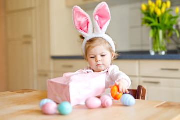 Cute little toddler girl wearing Easter bunny ears playing with colored pastel eggs. Happy baby child unpacking gifts. Adorable healthy smiling kid in pink clothes enjoying family holiday