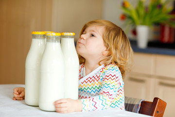 Wall Mural - Adorable toddler girl drinking cow milk for breakfast. Cute baby daughter with lots of bottles. Healthy child having milk as health calcium source. Kid at home or nursery in the morning.