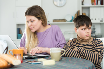 Young mother working at laptop and  unhappy preteen son