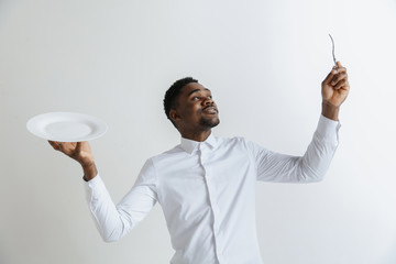 Young doubting attractive african american guy holding empty dish and fork isolated on grey background. Copy space and mock up. Blank template background.