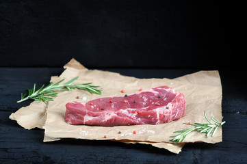 Wall Mural - Raw beef steak on paper. Rosemary, salt and pepper complement the composition. Dark wooden background. Close-up. 