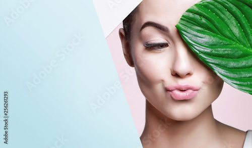 Cuadros en Lienzo Portrait of young beautiful woman with healthy glow perfect smooth skin holds green tropical leaf, look into the hole of colored paper