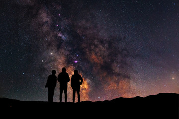 Wall Mural - three silhouettes stand on a hill and look at the close up milky way galaxy.