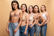 © LIGHTFIELD STUDIOS - selective focus of five beautiful multicultural girls in blue jeans and bras smiling and looking at camera isolated on beige, body positivity concept