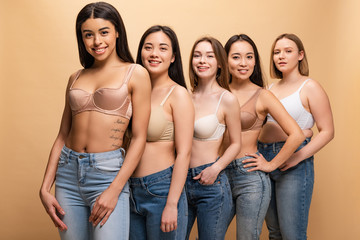 Wall Mural - selective focus of five beautiful multicultural girls in blue jeans and bras smiling and looking at camera isolated on beige, body positivity concept