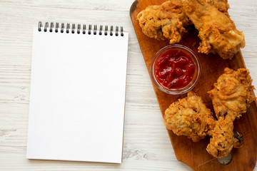 Wall Mural - Fried chicken drumsticks with sauce on wooden board, blank notepad over white wooden surface, overhead view. Flat lay, from above, top view.