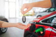 © estradaanton - Young family buying first electric car in the showroom. Close-up of male hand giving car key to female hand on battery electric car background. Eco car sale concept